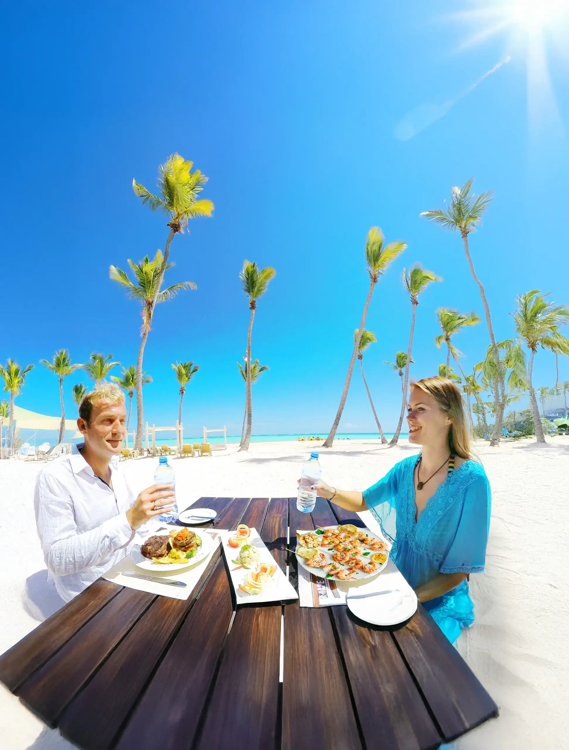 Pareja almorzando en una mesa de madera sobre la arena blanca, entre palmeras y bajo un cielo azul