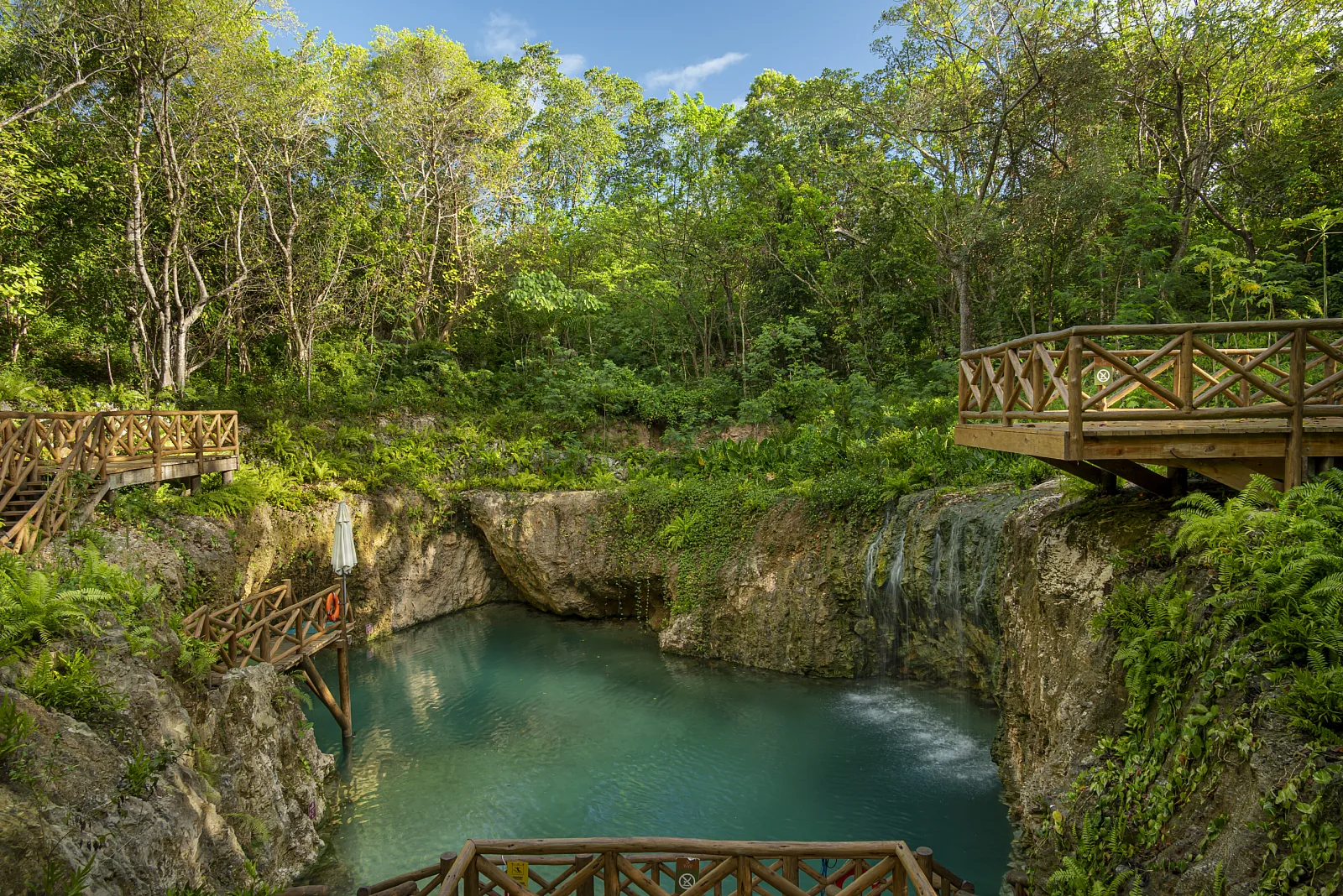 Cenote Hoyo Azul de Scape Park: piscina natural de agua turquesa rodeada de selva, con puentes y escaleras de madera