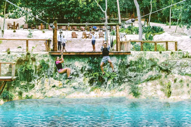 Dos personas cruzando en tirolina sobre el agua turquesa de un cenote, en plena selva